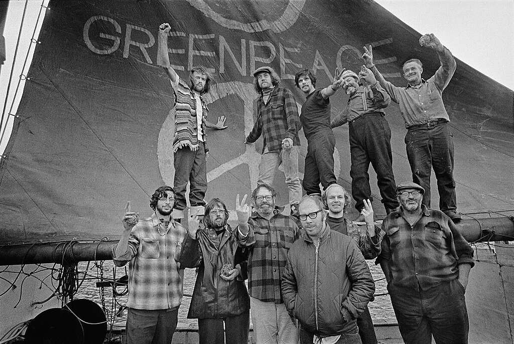 Black and white photograph of the crew from the first Greenpeace voyage making peace sign gestures with their hands onboard the Phyllis Cormack (also called "Greenpeace"). They are standing in-front of a large flag with the word “GREENPEACE” on it and a peace sign underneath. Caption: The crew of the Phyllis Cormack (also called "Greenpeace") on-board the ship. Clockwise from top left: Hunter, Moore, Cummings, Metcalfe, Birmingham, Cormack, Darnell, Simmons, Bohlen, Thurston, Fineberg. This is a photographic record by Robert Keziere of the very first Greenpeace voyage, which departed Vancouver on the 15th September 1971. The aim of the trip was to halt nuclear tests in Amchitka Island by sailing into the restricted area. Crew on-board the ship, are the pioneers of the green movement who formed the original group that became Greenpeace.