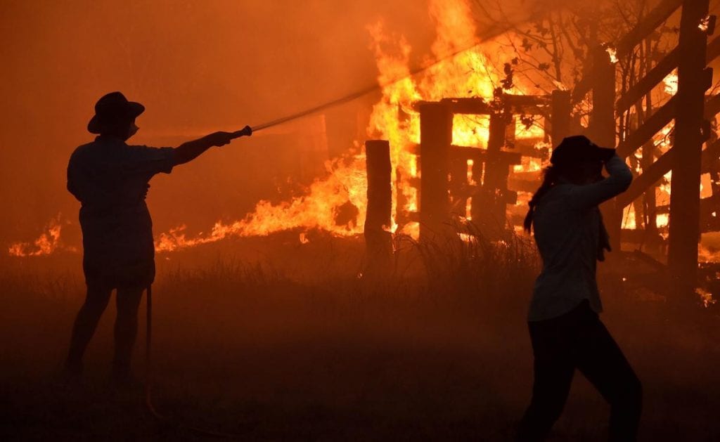 A farmer sprays water on a burning building from a garden hose in a field at night during a bushfire. 