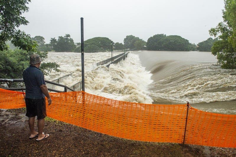 A man stands in front of an orange construction barrier beside the flooded Ross River. The river has risen over a pedestrian bridge and turbulence of the brown water is causing extreme damage.