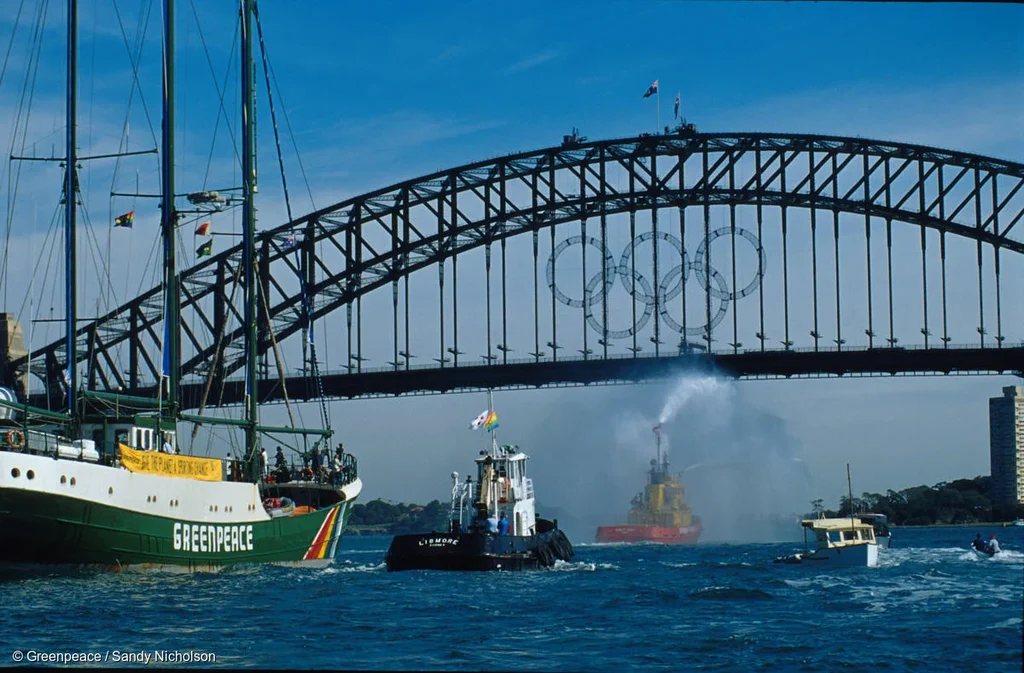 Rainbow Warrior at left side being towed by tugboat moving towards Sydney Harbour Bridge adorned with Olympic rings.